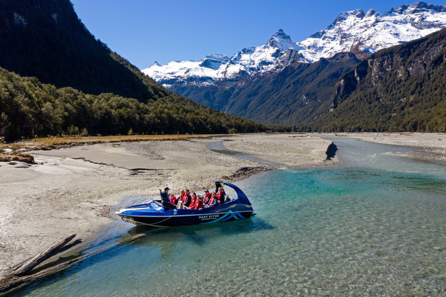 Wilderness Jet boat guide speaking to passengers on the Dart River, with snow-covered mountains behind them.