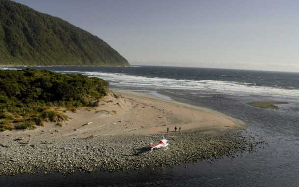 Aerial view of a helicopter on a rocky beach, with water and trees in the background