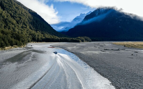 Aerial view of a jet boat travelling along the Dart River with funyak boats on the riverside, surrounded by mountain scenery.