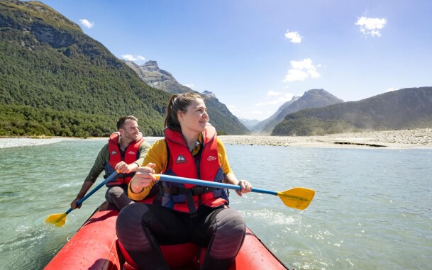 A woman and man paddling inflatable kayak on the Dart River, with native forest and mountains in the background.