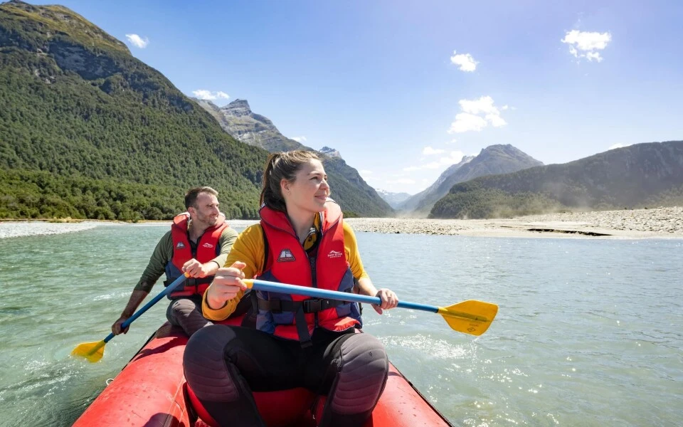A woman and man paddling inflatable kayak on the Dart River, with native forest and mountains in the background.