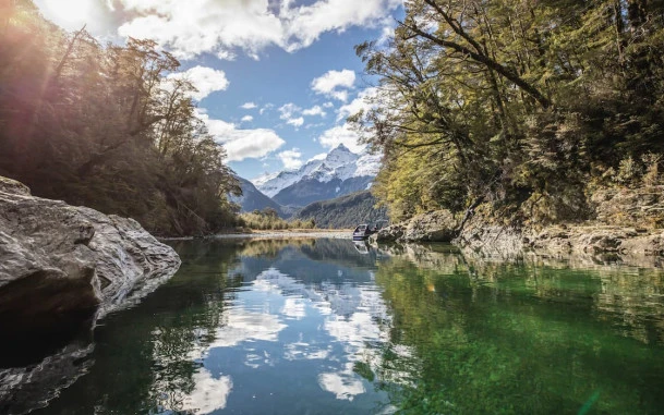 Still water on the Dart River reflecting surrounding trees, with mountains in the distance