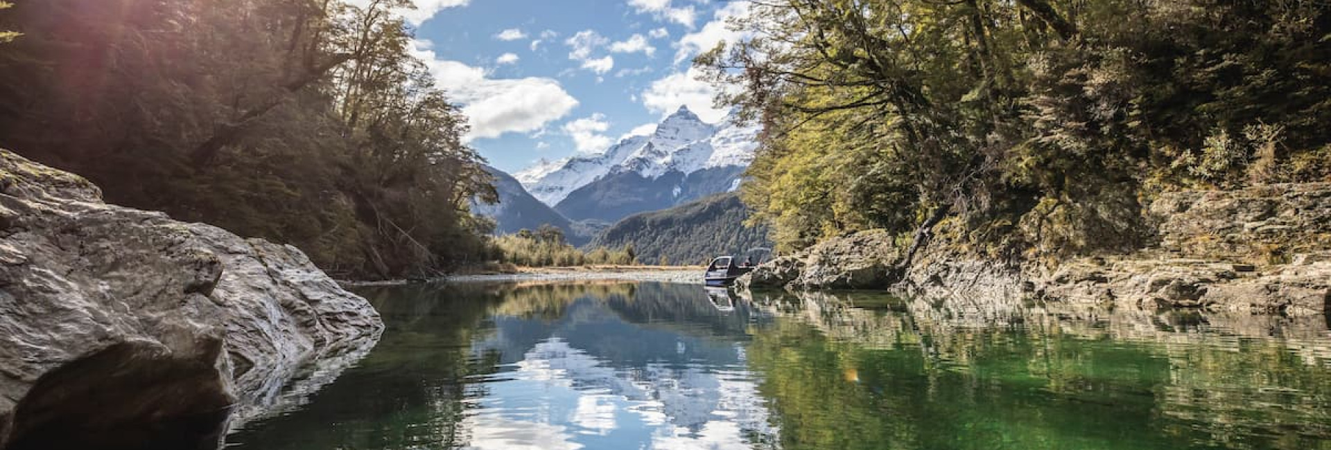 Still water on the Dart River reflecting surrounding trees, with mountains in the distance