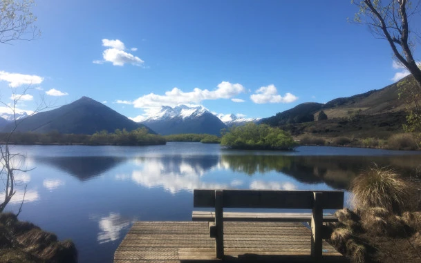 A wooden bench seat in Glenorchy facing the still water with mountains in the distance.