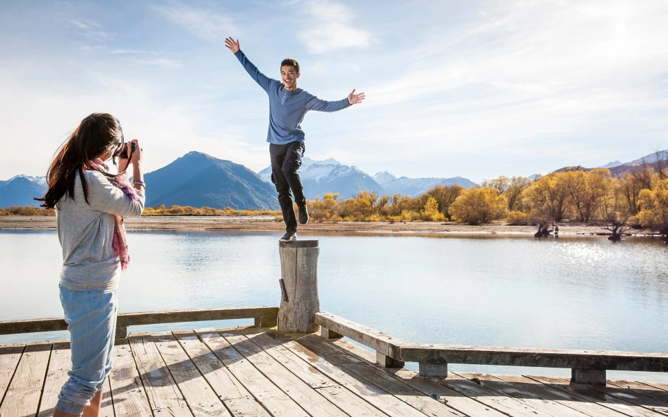 A man balancing on a pillar on the Glenorchy wharf as someone takes his photo, with mountains and water in the background.